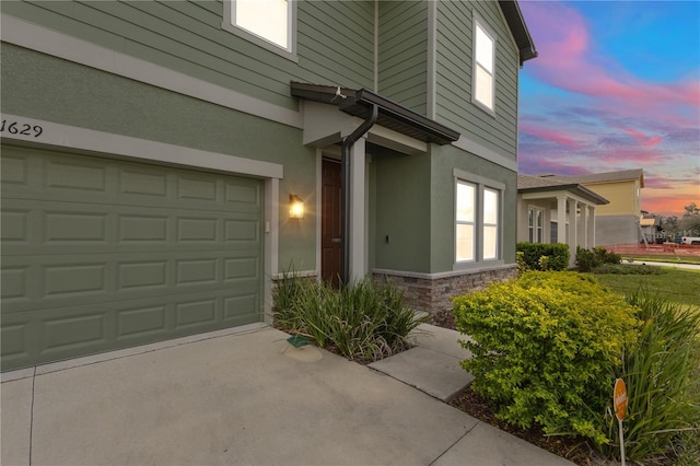 exterior entry at dusk featuring stone siding, an attached garage, concrete driveway, and stucco siding