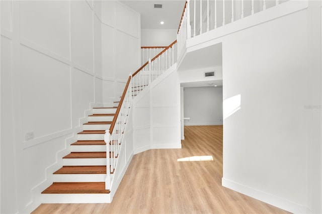 staircase with a decorative wall, wood finished floors, a towering ceiling, baseboards, and visible vents