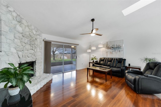 living room with vaulted ceiling with skylight, ceiling fan, wood finished floors, and a stone fireplace