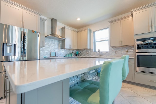 kitchen featuring wall chimney exhaust hood, light stone counters, light tile patterned floors, stainless steel appliances, and backsplash