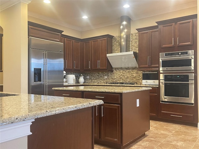 kitchen with a center island, backsplash, appliances with stainless steel finishes, light stone countertops, and wall chimney exhaust hood