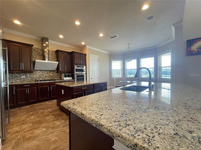 kitchen featuring visible vents, a kitchen island with sink, a sink, wall chimney range hood, and gas cooktop