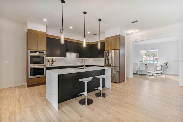 kitchen with stainless steel appliances, light hardwood / wood-style floors, a kitchen island with sink, and hanging light fixtures