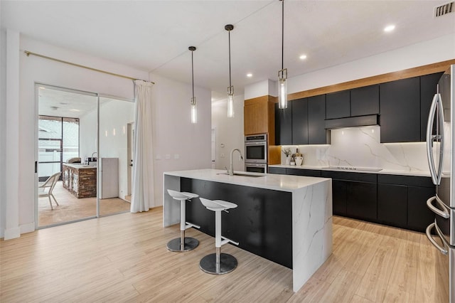 kitchen featuring a kitchen island with sink, hanging light fixtures, backsplash, stainless steel appliances, and light wood-type flooring