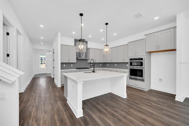 kitchen featuring gas stovetop, dark hardwood / wood-style floors, gray cabinets, stainless steel double oven, and a kitchen island with sink