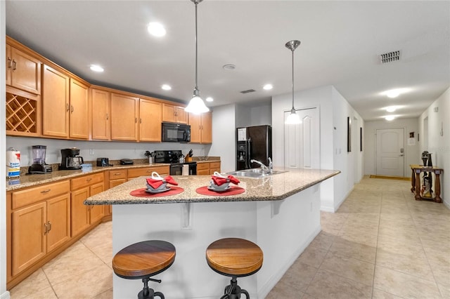 kitchen with sink, hanging light fixtures, light stone counters, black appliances, and a center island with sink