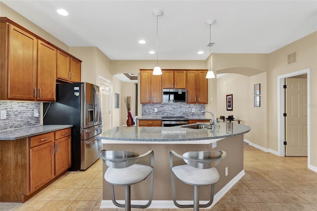 kitchen with an island with sink, stainless steel appliances, sink, light stone counters, and pendant lighting