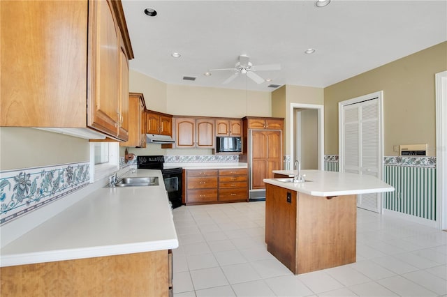 kitchen with brown cabinets, light countertops, an island with sink, under cabinet range hood, and black appliances