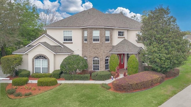 view of front facade featuring a shingled roof, a front yard, brick siding, and stucco siding