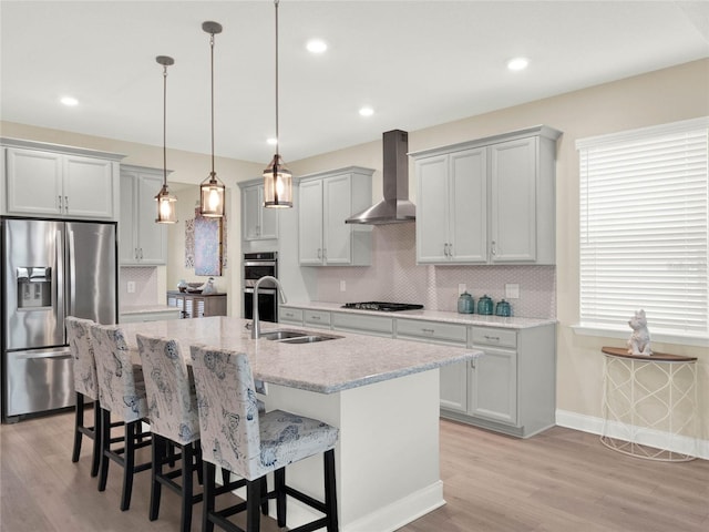 kitchen featuring decorative light fixtures, a center island with sink, appliances with stainless steel finishes, a sink, and wall chimney range hood