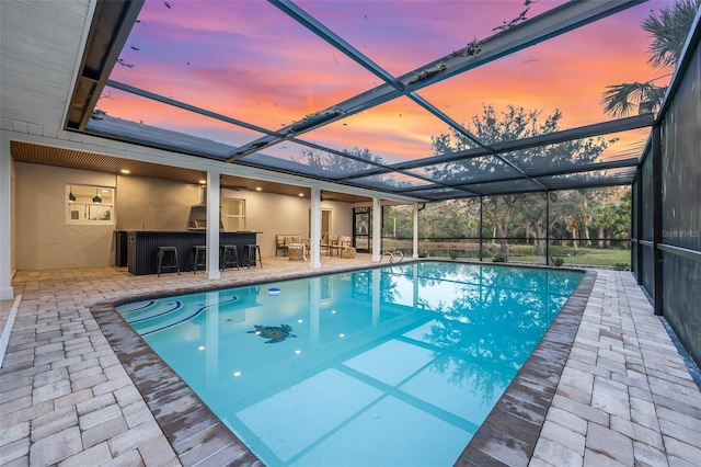 pool at dusk with a patio, glass enclosure, and an outdoor bar