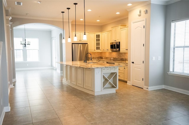 kitchen with decorative light fixtures, glass insert cabinets, appliances with stainless steel finishes, an island with sink, and light stone countertops