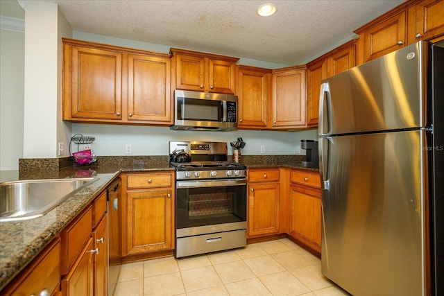 kitchen with a textured ceiling, light tile patterned flooring, appliances with stainless steel finishes, brown cabinets, and dark stone counters