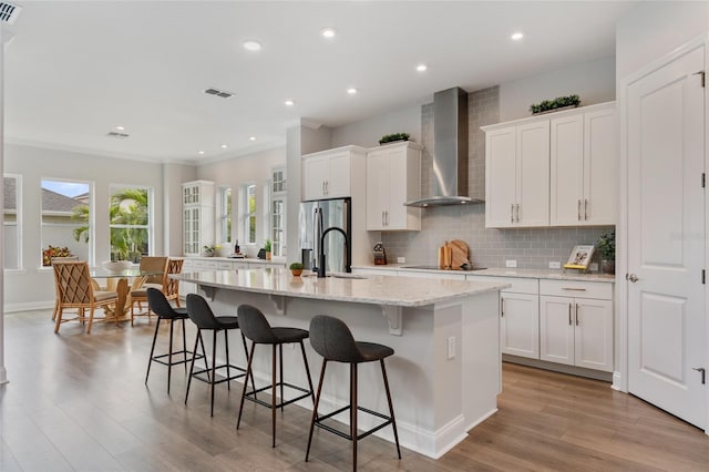 kitchen with visible vents, backsplash, stainless steel fridge with ice dispenser, wall chimney exhaust hood, and an island with sink