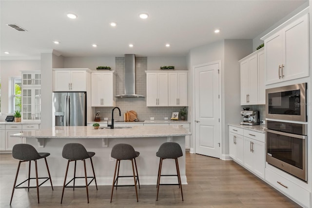 kitchen featuring a center island with sink, appliances with stainless steel finishes, white cabinetry, a sink, and wall chimney exhaust hood