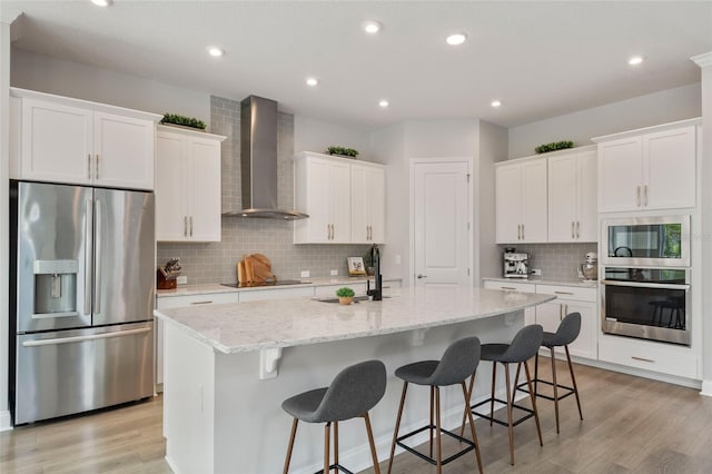 kitchen with a kitchen island with sink, stainless steel appliances, light wood-type flooring, wall chimney range hood, and white cabinetry