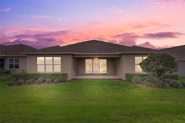 back of house at dusk featuring roof with shingles, a lawn, a patio, and stucco siding