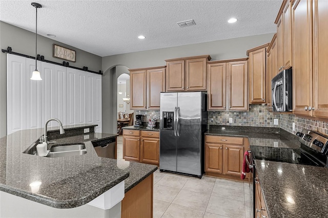 kitchen featuring a barn door, a center island with sink, appliances with stainless steel finishes, decorative light fixtures, and a sink