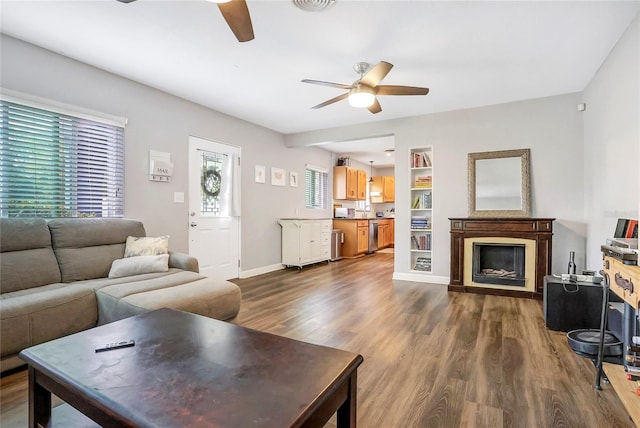 living room with baseboards, dark wood-style floors, ceiling fan, built in shelves, and a fireplace