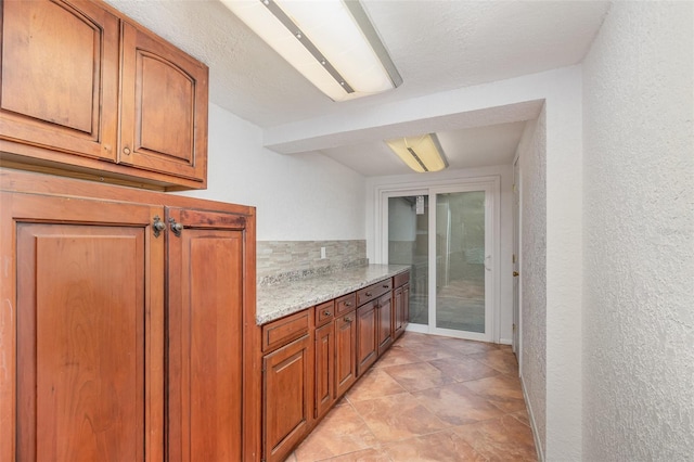 kitchen featuring light stone countertops, brown cabinets, backsplash, and a textured wall