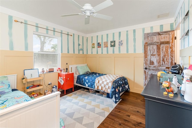 bedroom with wallpapered walls, visible vents, a wainscoted wall, ceiling fan, and wood finished floors