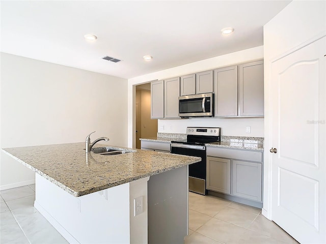 kitchen with light stone counters, stainless steel appliances, a sink, gray cabinets, and an island with sink