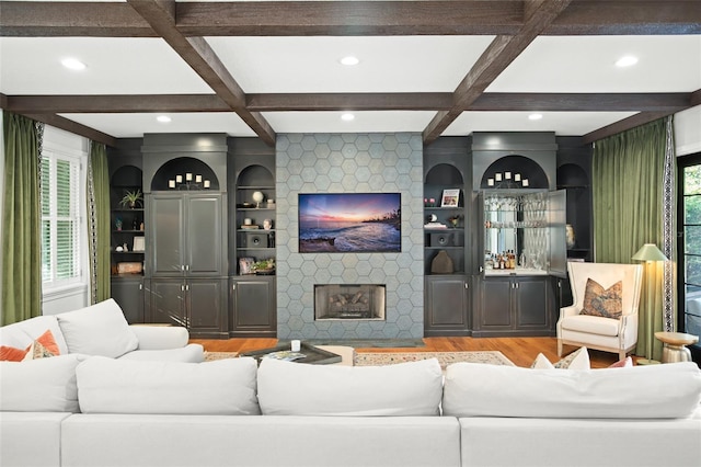 living area with beam ceiling, coffered ceiling, a stone fireplace, and wood finished floors