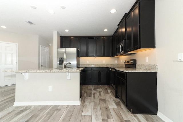 kitchen featuring appliances with stainless steel finishes, dark cabinets, light stone counters, and an island with sink