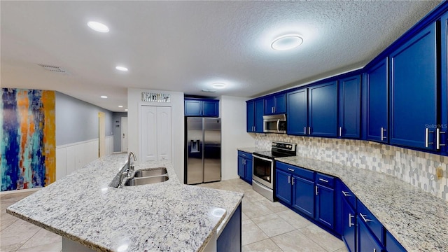 kitchen with a kitchen island with sink, stainless steel appliances, a sink, blue cabinetry, and light stone countertops