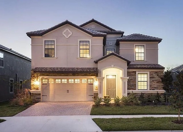 mediterranean / spanish-style house featuring a garage, a tile roof, stone siding, decorative driveway, and stucco siding