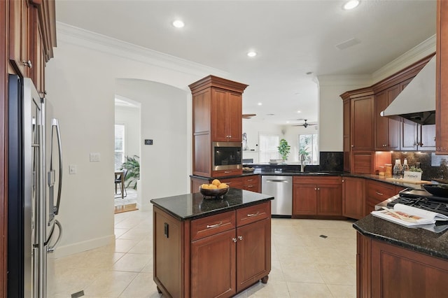 kitchen featuring arched walkways, appliances with stainless steel finishes, wall chimney range hood, dark stone counters, and a peninsula