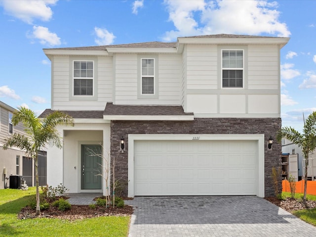 view of front of property with an attached garage, central AC, a shingled roof, stone siding, and decorative driveway