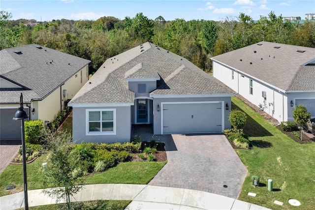 view of front of house with decorative driveway, a shingled roof, a wooded view, a garage, and a front lawn