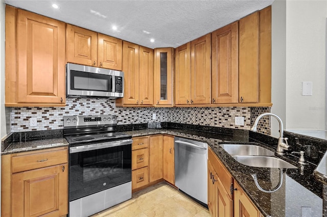 kitchen with brown cabinets, appliances with stainless steel finishes, glass insert cabinets, a sink, and dark stone counters