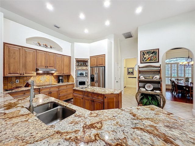 kitchen with brown cabinetry, appliances with stainless steel finishes, light stone countertops, under cabinet range hood, and a sink