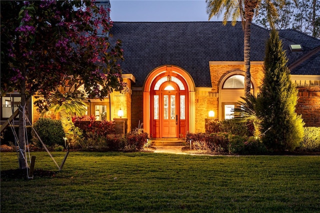view of exterior entry featuring brick siding, a lawn, and a shingled roof