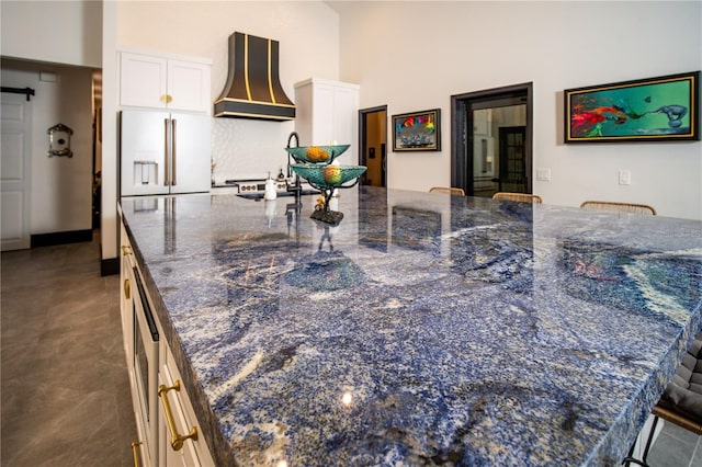 kitchen featuring dark stone countertops, high end fridge, a towering ceiling, custom range hood, and white cabinets