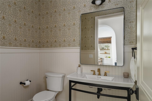 bathroom with wainscoting, a sink, toilet, and wallpapered walls