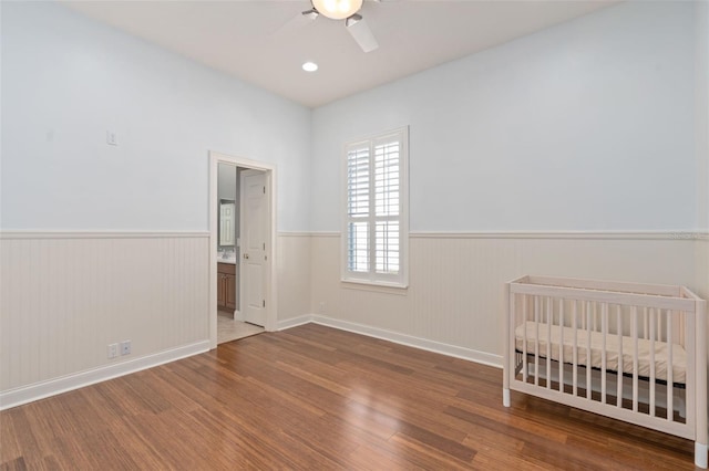 unfurnished bedroom featuring a nursery area, a ceiling fan, wood finished floors, and wainscoting