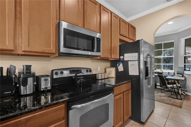 kitchen with light tile patterned flooring, appliances with stainless steel finishes, brown cabinets, dark stone counters, and crown molding