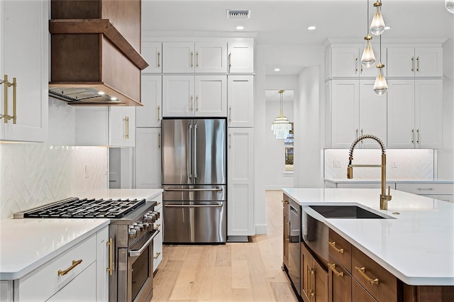 kitchen featuring a kitchen island with sink, a sink, white cabinetry, hanging light fixtures, and appliances with stainless steel finishes