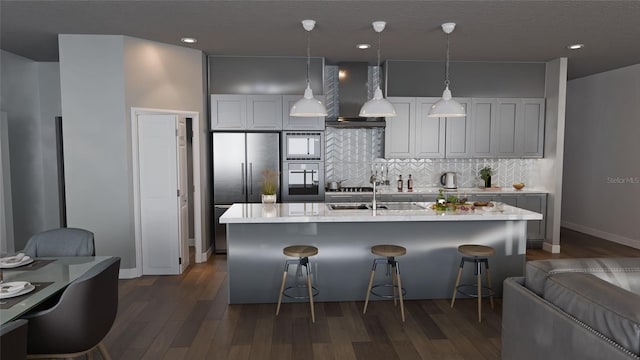 kitchen featuring dark wood-style flooring, light countertops, wall chimney exhaust hood, a center island with sink, and pendant lighting