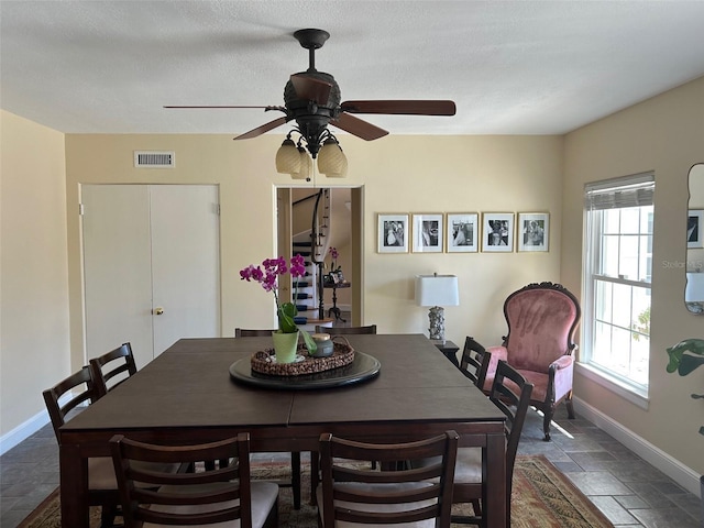 dining room featuring stone tile floors, visible vents, ceiling fan, a textured ceiling, and baseboards