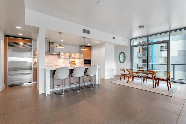 kitchen with light countertops, visible vents, wall chimney range hood, built in appliances, and a kitchen bar