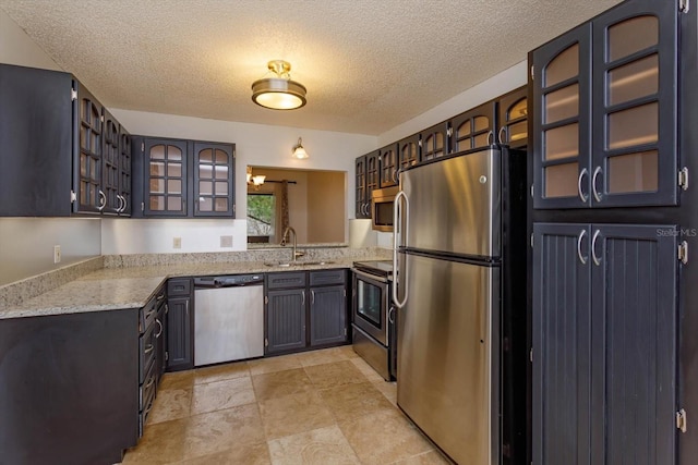 kitchen with a textured ceiling, light stone counters, a sink, appliances with stainless steel finishes, and glass insert cabinets