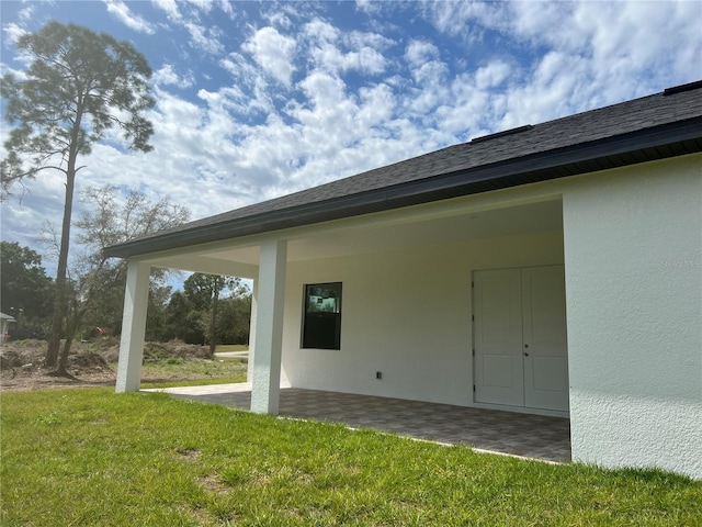 exterior space with roof with shingles, a patio, a lawn, and stucco siding