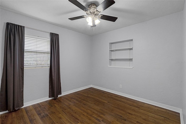 spare room with dark wood-type flooring, built in shelves, baseboards, and a ceiling fan