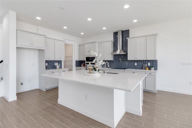 kitchen with a sink, an island with sink, light countertops, and wall chimney range hood