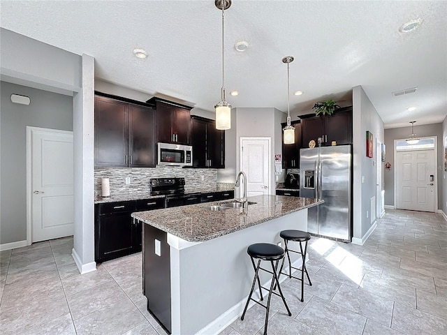 kitchen featuring visible vents, dark stone counters, an island with sink, appliances with stainless steel finishes, and a sink