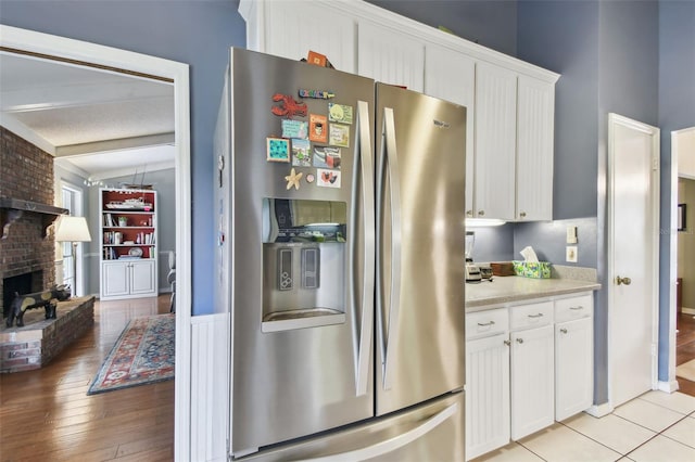 kitchen with stainless steel refrigerator with ice dispenser, light tile patterned floors, light countertops, a brick fireplace, and white cabinetry